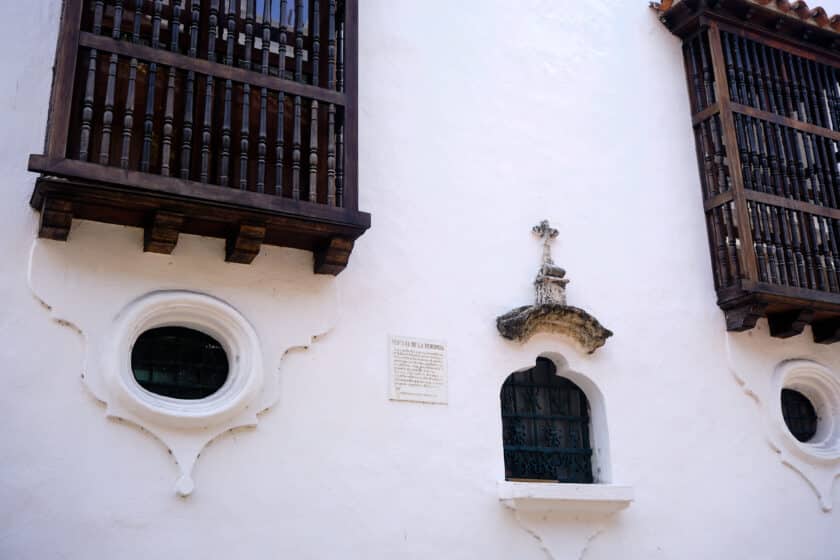 A small window in a white wall with larger wooden windows at the Palacio de la Inquisición Cartagena.