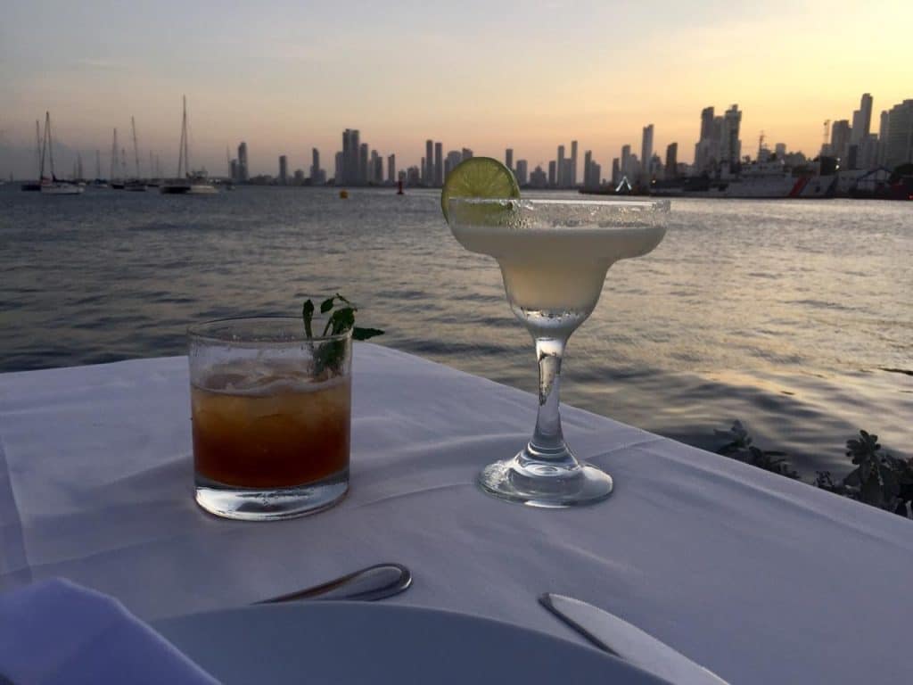 A photo of two drinks on a table with the bay of Cartagena beyond and the sky lit up with the colors of sunset taken from Club de Pesca, on our list of places to get sunset drinks in Cartagena.