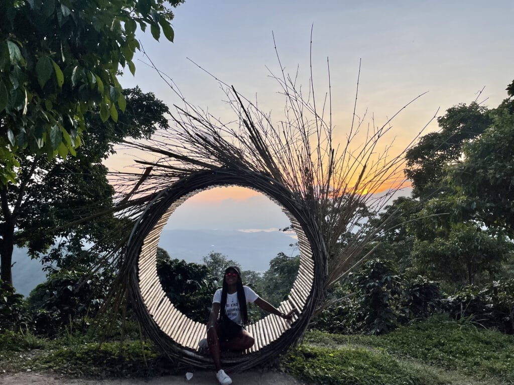 Photo of a girl sitting in a circle wooden circle with mountains and the sunset behind her.