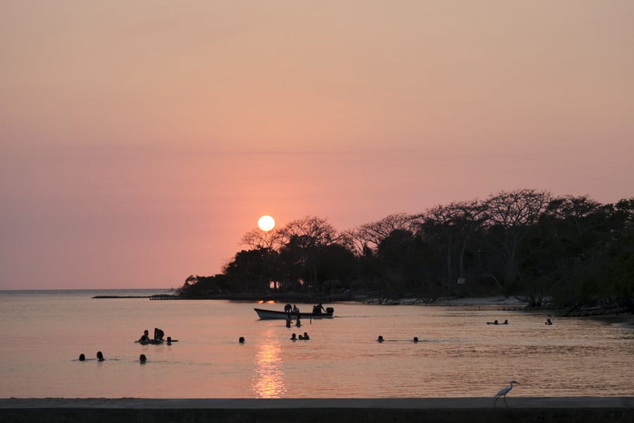 People swimming in the water in front a hotel in the Rosario Islands with sunset in the background over some trees.