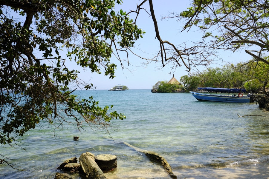 Photo of clear blue waters seen through tree branches with a small empty boat, and a small island house in the background.