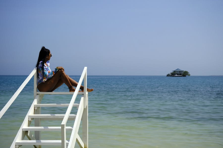 Photo of a girl in a bathing suit sitting on the top of a short white stairwell in the water in the Rosario Islands with a tiny island with a house in the background.