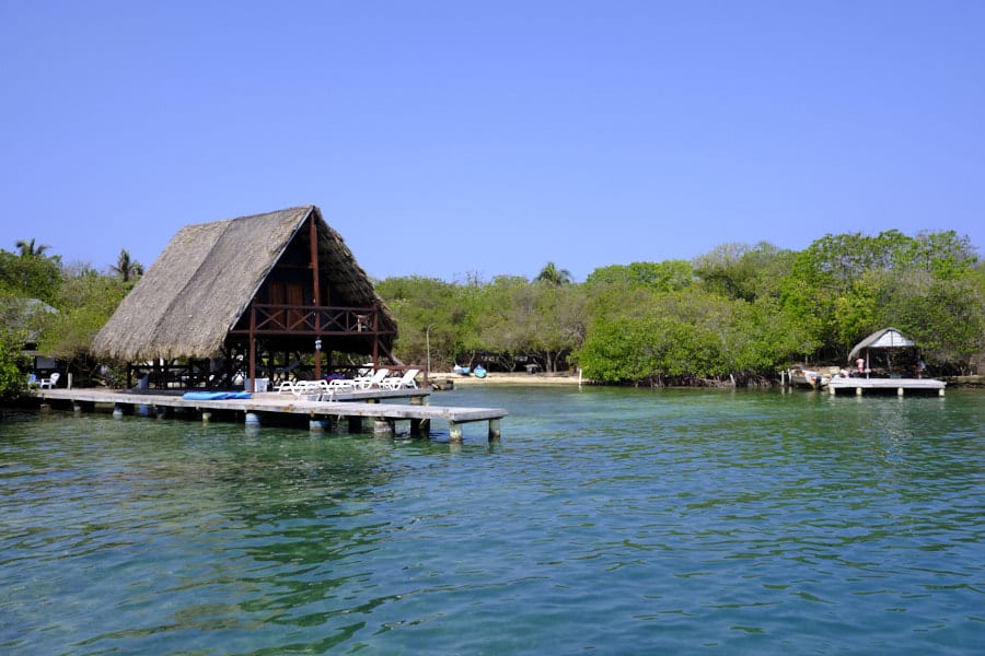 Photo of a house with a thatch roof with a deck over the water in the Rosario Islands with a small beach and trees in the background.