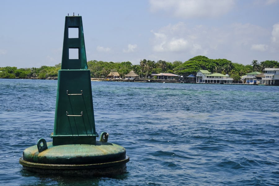 Photo of a green floating buoy in the Rosario Islands near Cartagena with houses on the shore behind it.