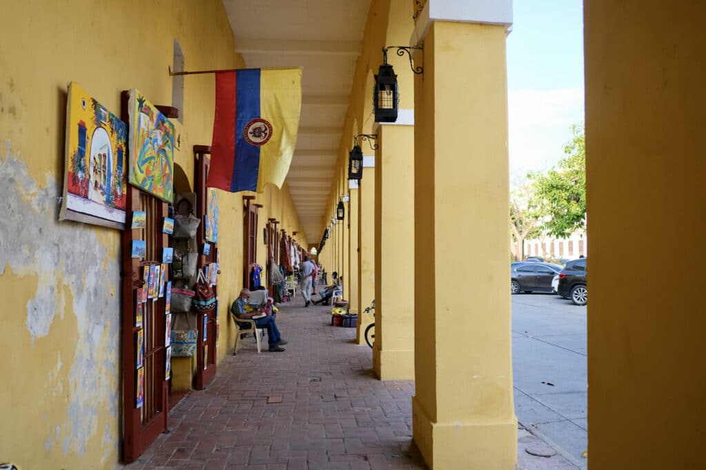 Narrow passage way under yellow columns with a Colombian flag and art hanging from the wall and people sitting and walking in the background.