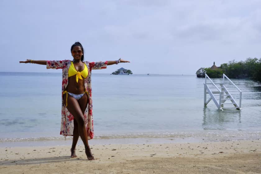 Girl in a robe and bikini smiling with her arms raised to show the best Rosario Island day trip with a small white staircase in the water and a small house on a island in the background.