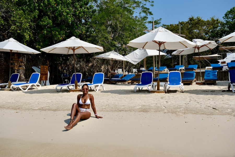 A girl in a bikini sitting on a beach during a day trip to Rosario Islands from Cartagena with blue beach chairs and umbrellas behind her.