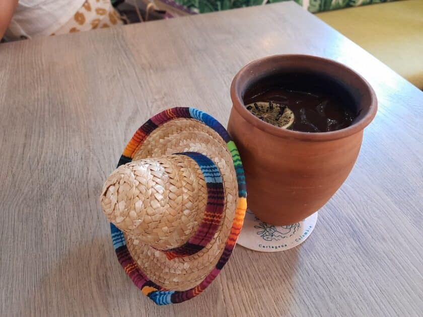 Photo of a clay cup with a smoky lime  inside and a small straw sombrero with colorful stripes on the table next to it.
