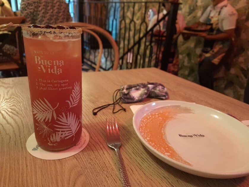 Photo of red liquid in a tall glass that says Buena Vida next to a plate on a table that also says Buena Vida.