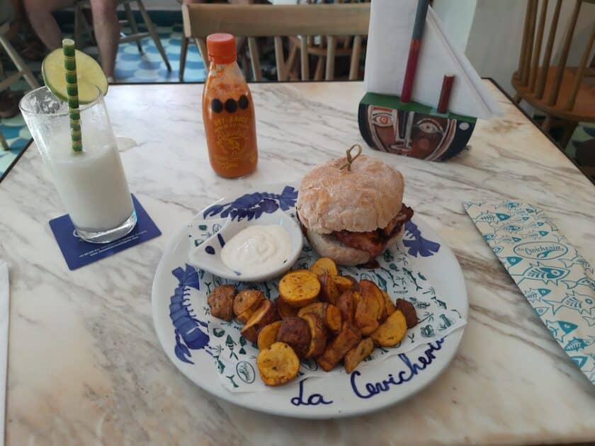 Photo of a table with a plate that says cevichería with a sandwich and potatoes, a drink, hot sauce, and napkins also on the table.