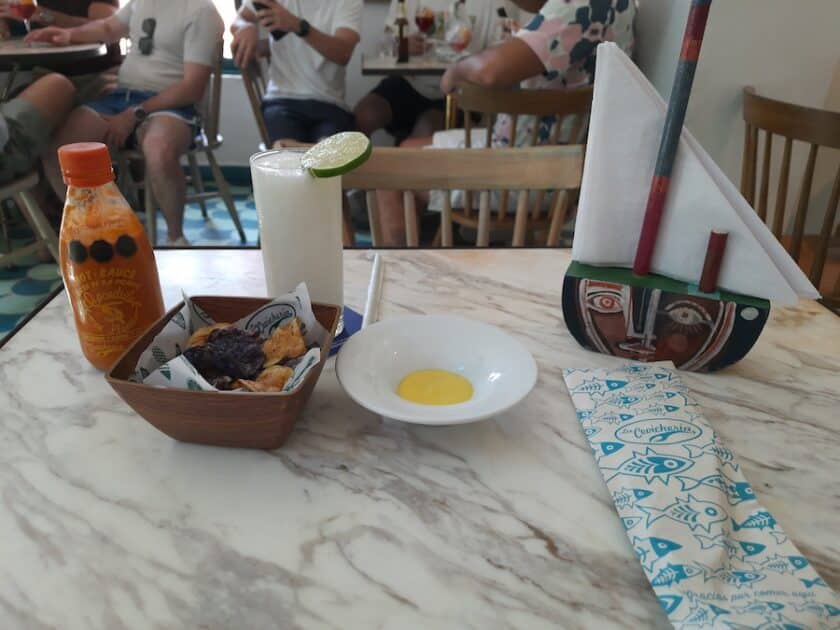 Photo of a table at La Cevicheria Restaurant in Cartagena with a small bowl of chips, bowl of yellow sauce, a hot sauce bottle, and napkin holder that looks like a sail boat.