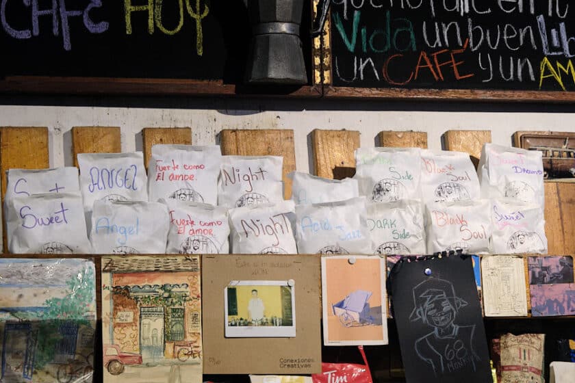 Photo of white bags of coffee with colorful names on shelf above post cards and drawings.
