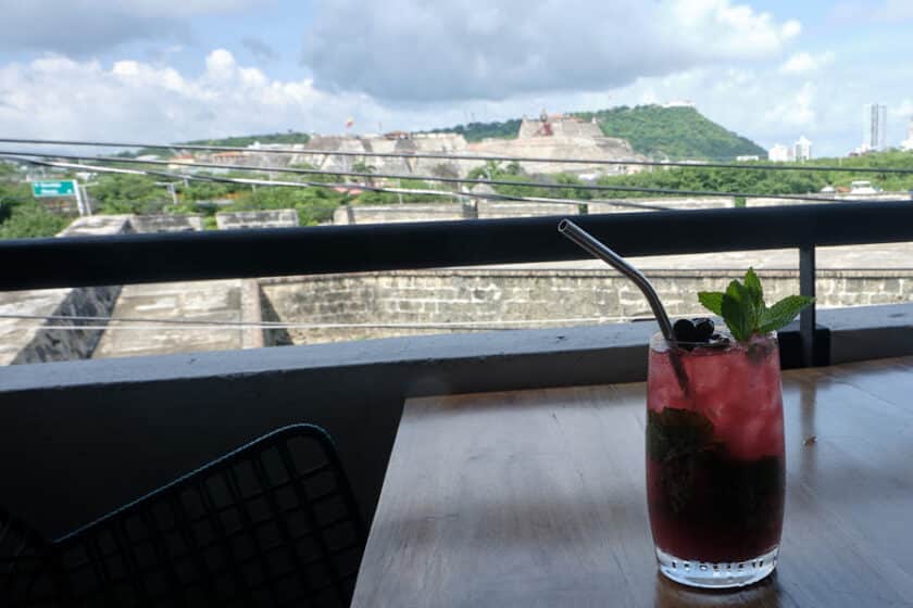 A glass with a drink with a mint garnish and straw on a table with the Cartagena fort in the background.