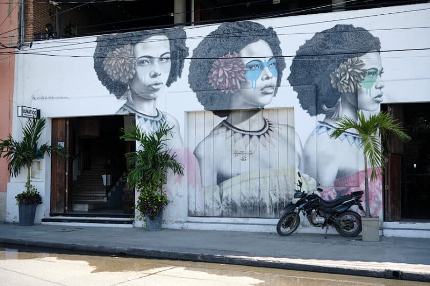 Outside of Lunático with a mural showing the same woman 3 times with different colored flowers and face paint and a motorcycle parked in front.