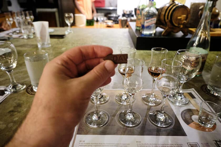 Hand holding a small piece of chocolate with tasting glasses on a table in the background during a rum and chocolate tasting in Cartagena.