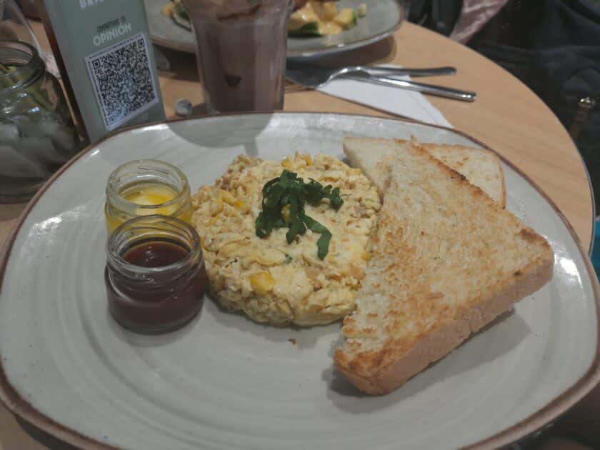 Plate with scrambled eggs, toast, and tiny jars of butter and jam.